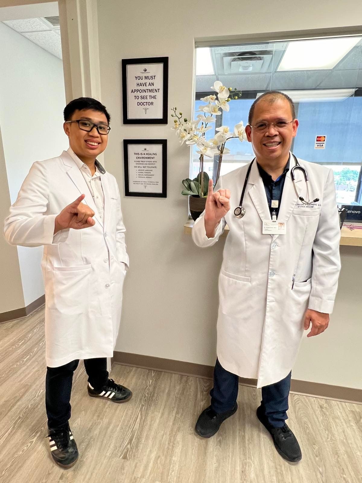 Two individuals wearing white lab coats, standing in a medical office near signs and an orchid plant.