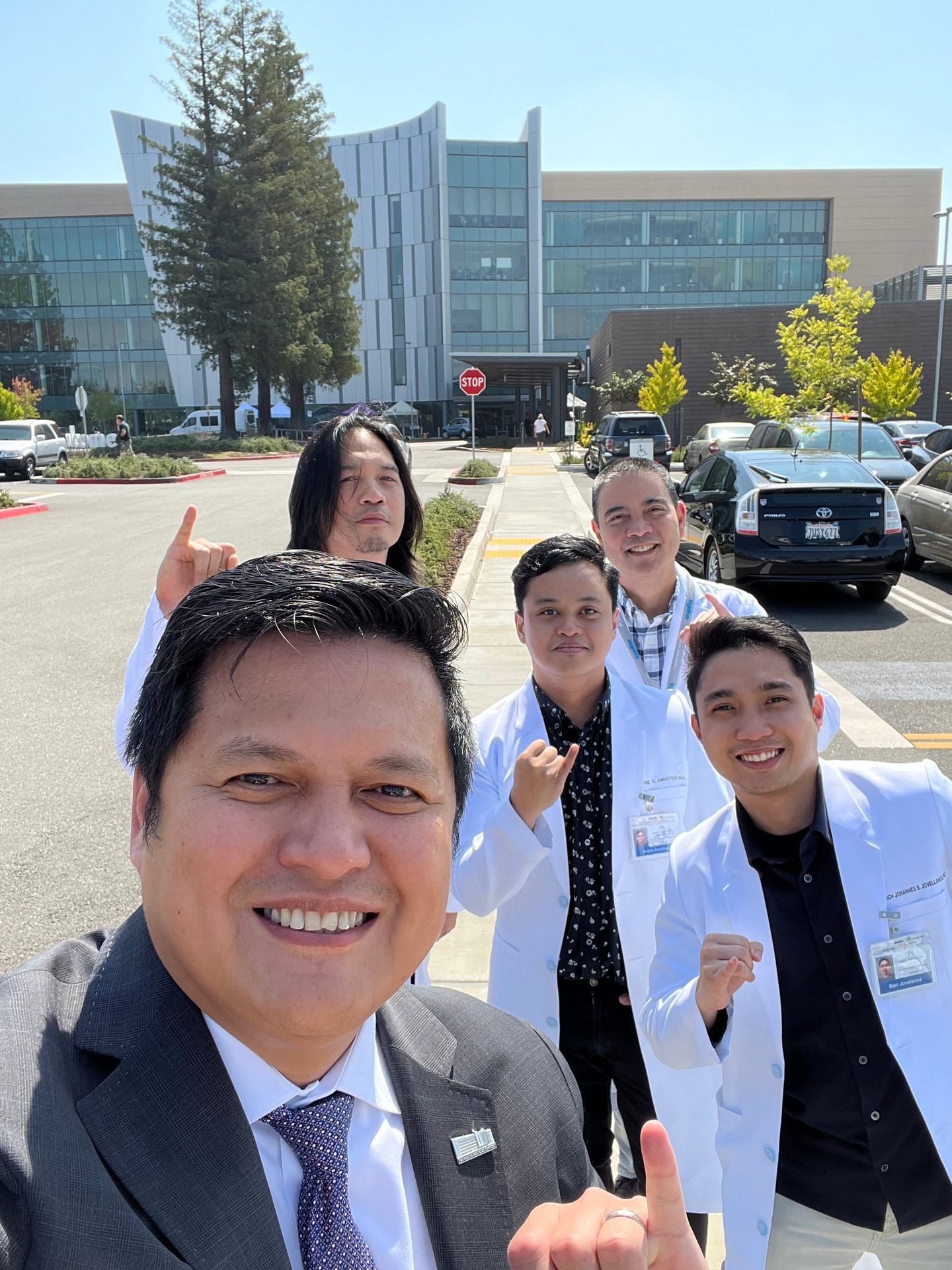 Group of smiling people, some in lab coats, taking a selfie outside a modern building.