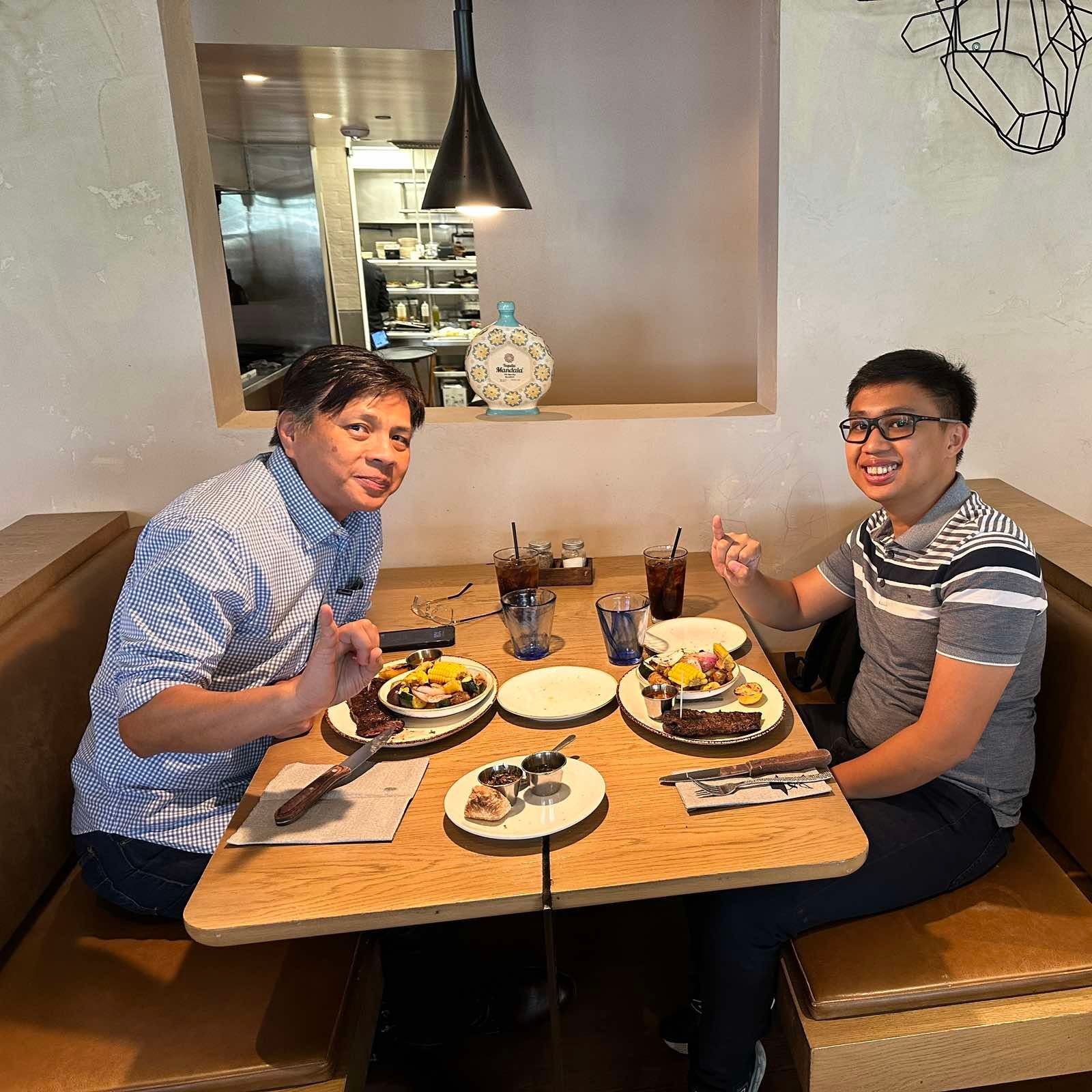 Two people seated at a restaurant booth, enjoying meals with drinks on a wooden table.