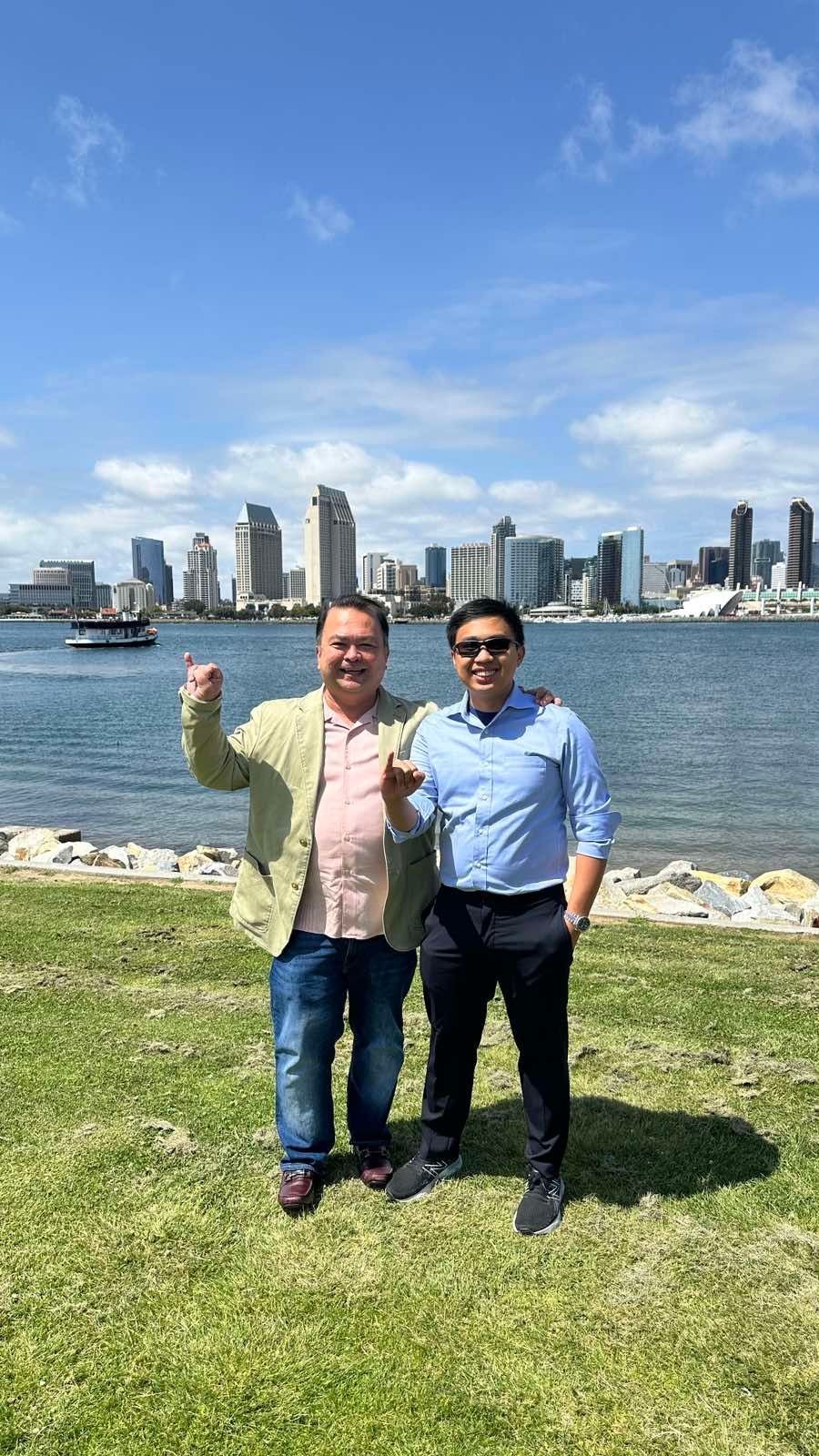 Two people standing on grass near water with a city skyline and a boat in the background.