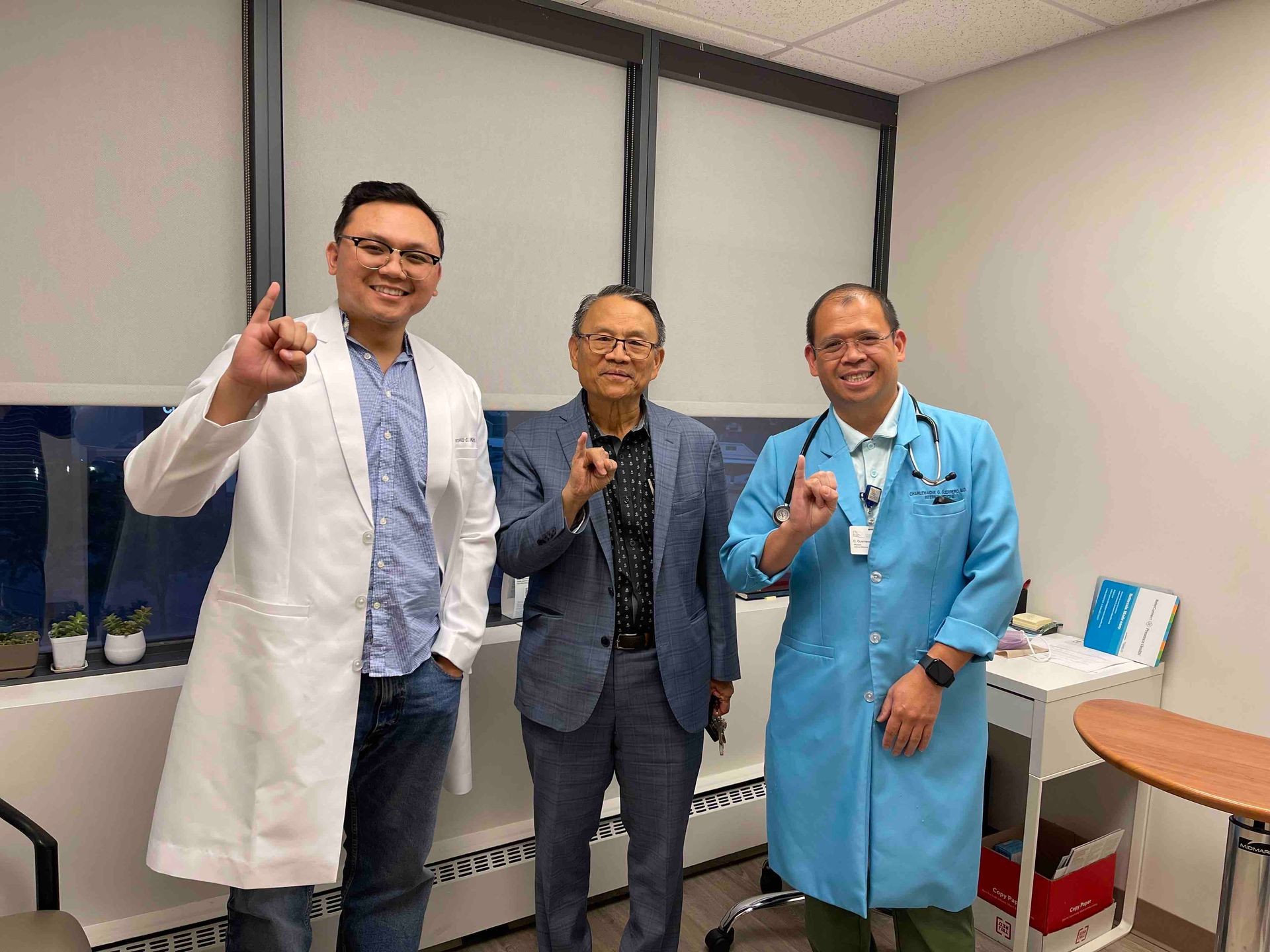 Three men smiling in an office, wearing lab coats and gesturing with index fingers raised.