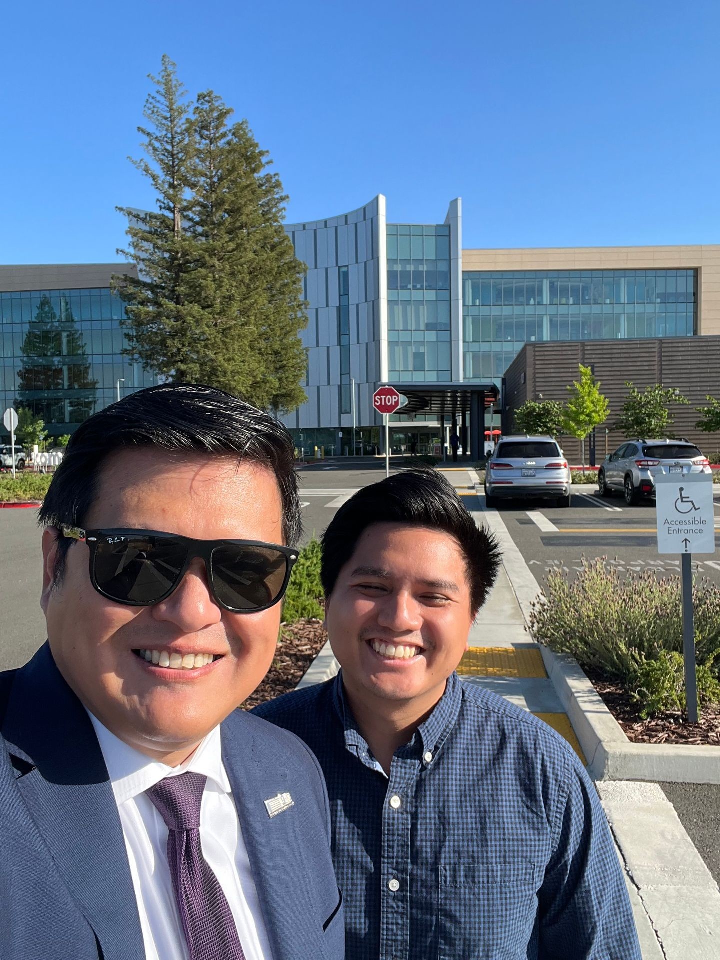 Two men smiling in front of a modern glass building with a clear blue sky.