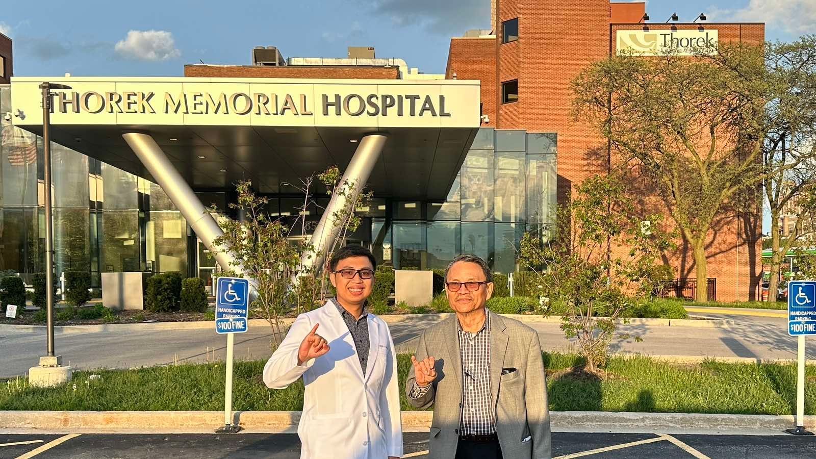 Two men posing outside Thorek Memorial Hospital with a glass entrance and brick building in the background.